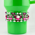 A collection of beaded bracelets in shades of pink, green, white, and silver. The bracelets are stacked on a bright green tumbler. The beads vary in size and material, creating a layered and textured look. The bracelets are a mix of round and flat beads.
