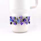A floral bracelet with purple and blue flower beads, clear beads, and silver accents. The bracelet is wrapped around a white mug. The flowers have green leaves.