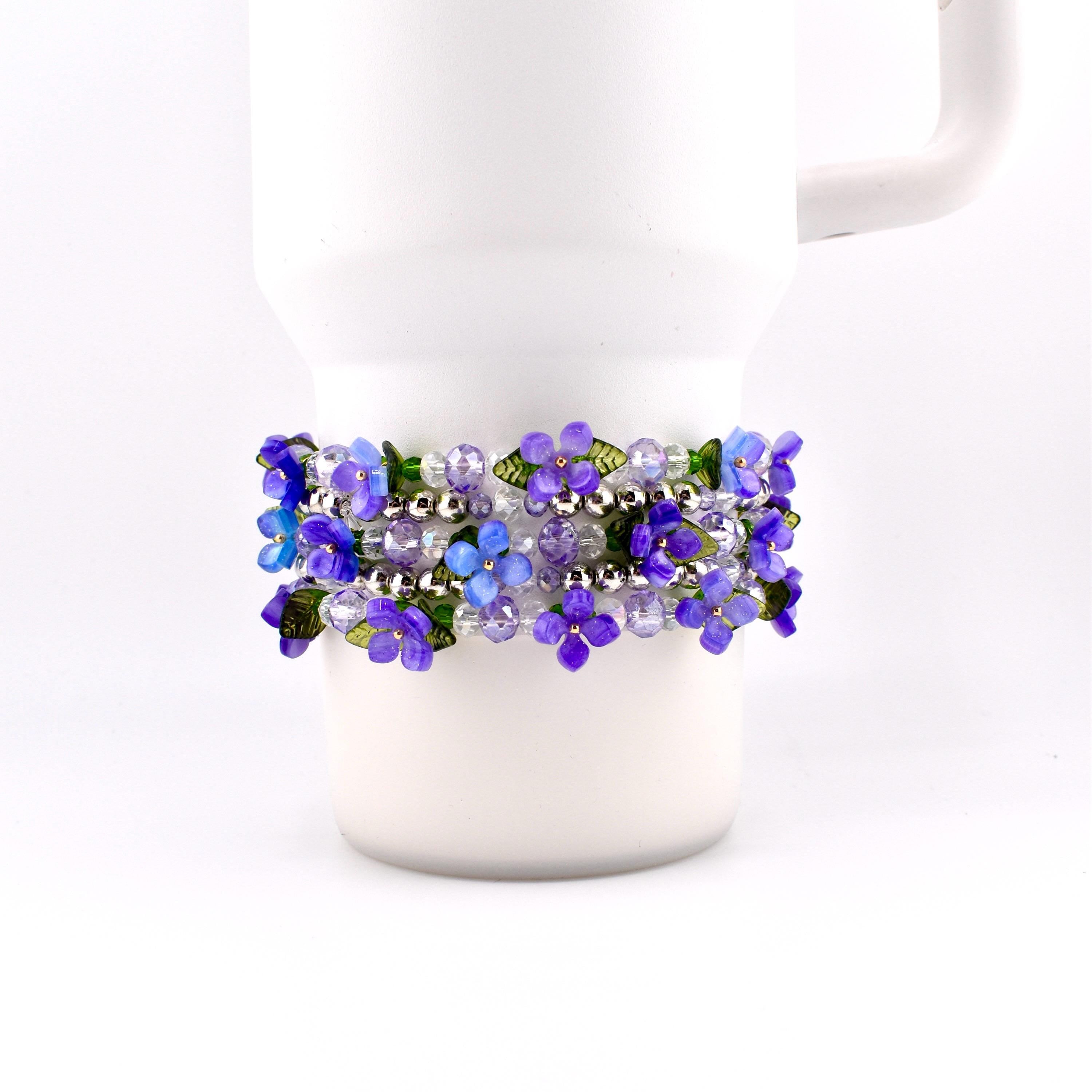 A floral bracelet with purple and blue flower beads, clear beads, and silver accents. The bracelet is wrapped around a white mug. The flowers have green leaves.