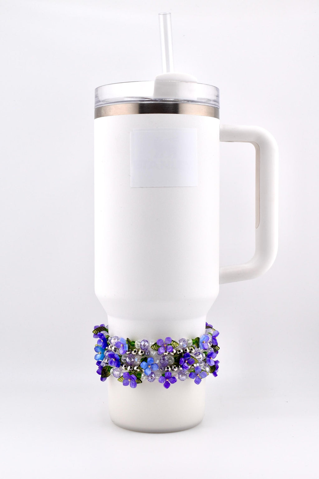 White insulated tumbler with a handle, clear lid, and straw. A decorative beaded band with purple and blue flowers encircles the base of the tumbler.