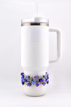 White insulated tumbler with a handle, clear lid, and straw. A decorative beaded band with purple and blue flowers encircles the base of the tumbler.