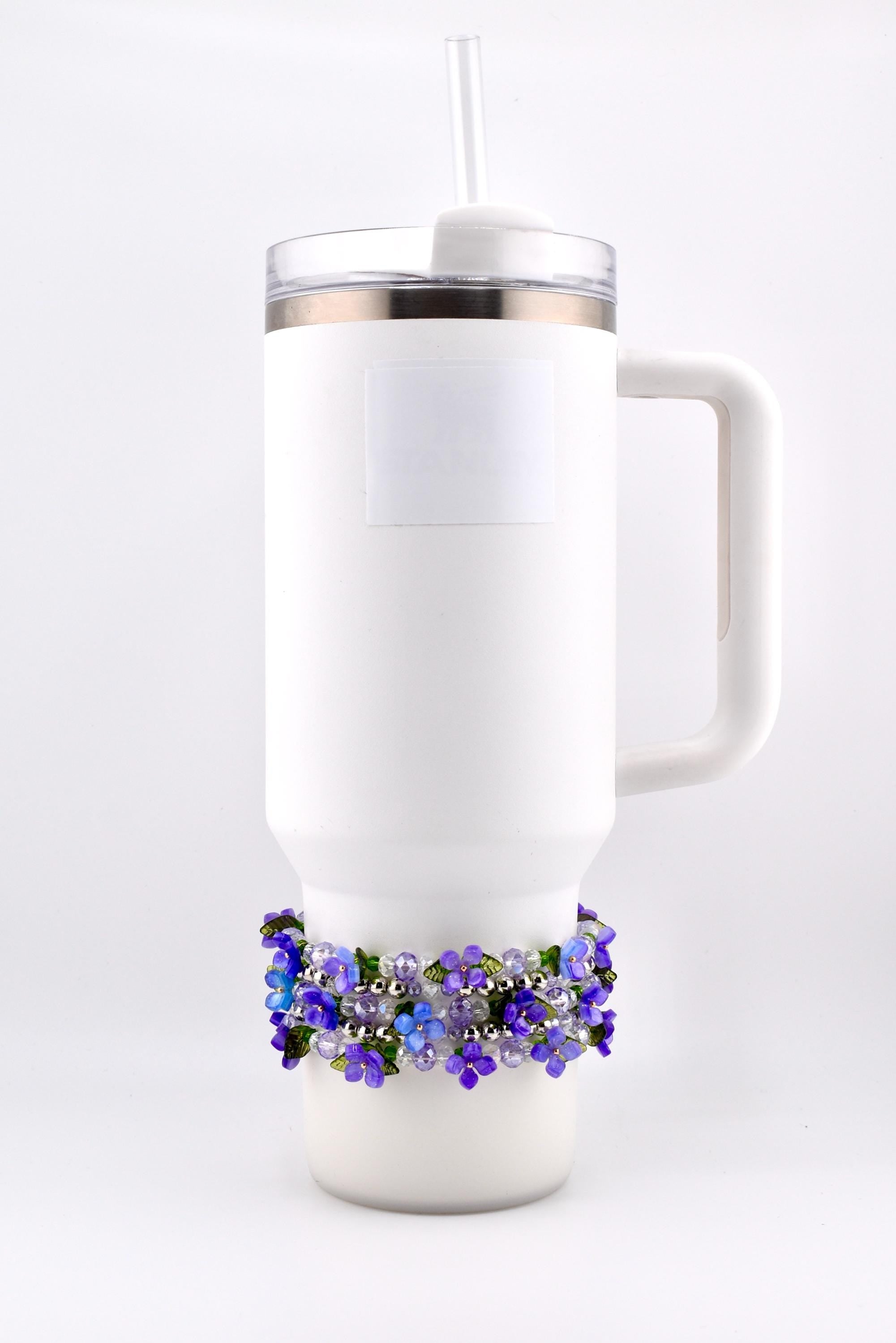 White insulated tumbler with a handle, clear lid, and straw. A decorative beaded band with purple and blue flowers encircles the base of the tumbler.