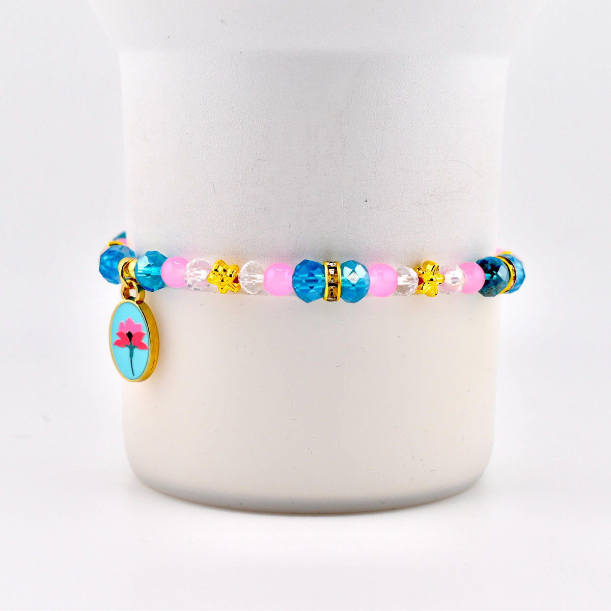 A beaded bracelet featuring a floral charm. The bracelet has blue, pink, and clear beads, with gold-colored accents. The charm is an oval shape with a pink flower on a blue background.