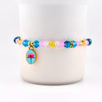 A beaded bracelet featuring a pink and blue color scheme. The bracelet has blue and pink beads, with gold accents and a charm with a pink flower on a blue background. The bracelet is displayed on a white background.