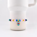A beaded bracelet with a floral charm. The bracelet features pink, blue, yellow, and clear beads. The charm is an oval shape with a blue background and a red flower design. The bracelet is displayed on a white mug.