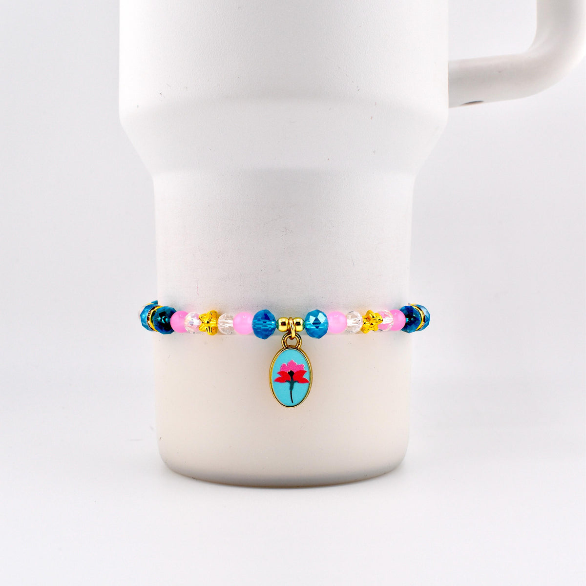 A beaded bracelet with a floral charm. The bracelet features pink, blue, yellow, and clear beads. The charm is an oval shape with a blue background and a red flower design. The bracelet is displayed on a white mug.