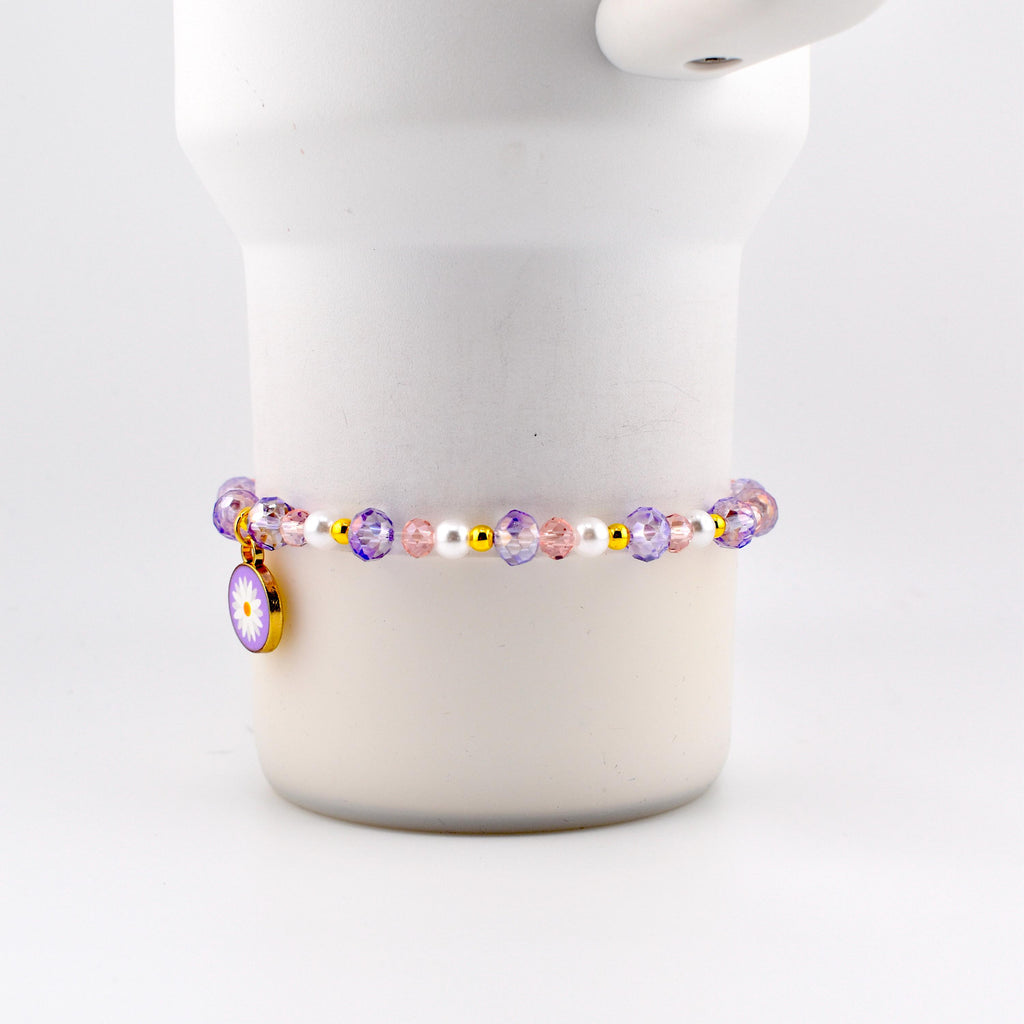 A beaded bracelet featuring a daisy charm. The bracelet has a mix of purple, pink, white, and gold beads. The daisy charm is gold with a purple and white flower design.