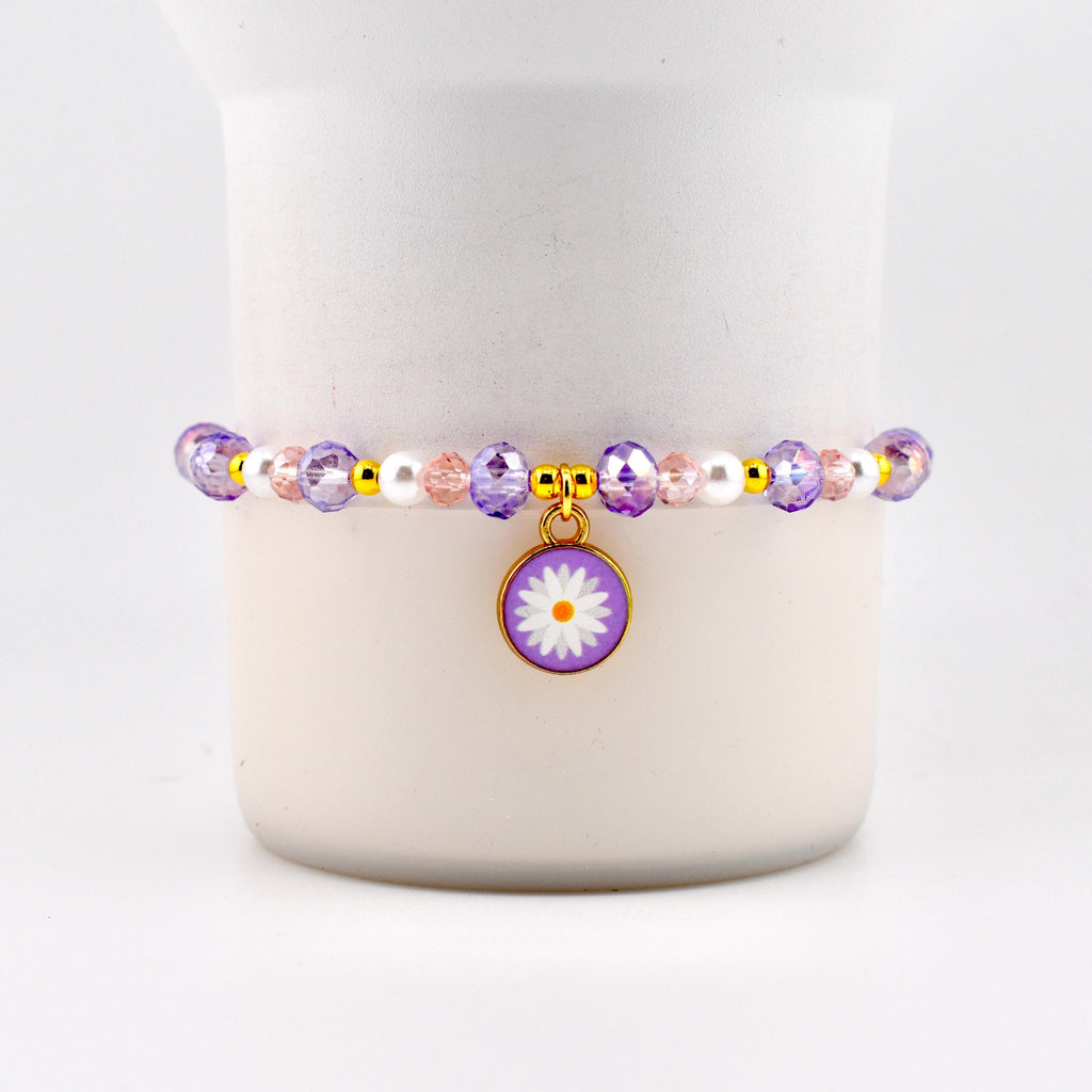 A beaded bracelet featuring a daisy charm. The bracelet has purple, pink, and white beads with gold accents. The daisy charm is purple with a white flower and a gold border. A delicate piece of jewelry.