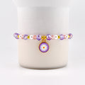 A beaded bracelet featuring a daisy charm. The bracelet has purple, pink, and white beads with gold accents. The daisy charm is purple with a white flower and a gold border. A delicate piece of jewelry.