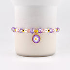A beaded bracelet featuring a daisy charm. The bracelet has purple, pink, and white beads with gold accents. The daisy charm is purple with a white flower and a gold border. A delicate piece of jewelry.
