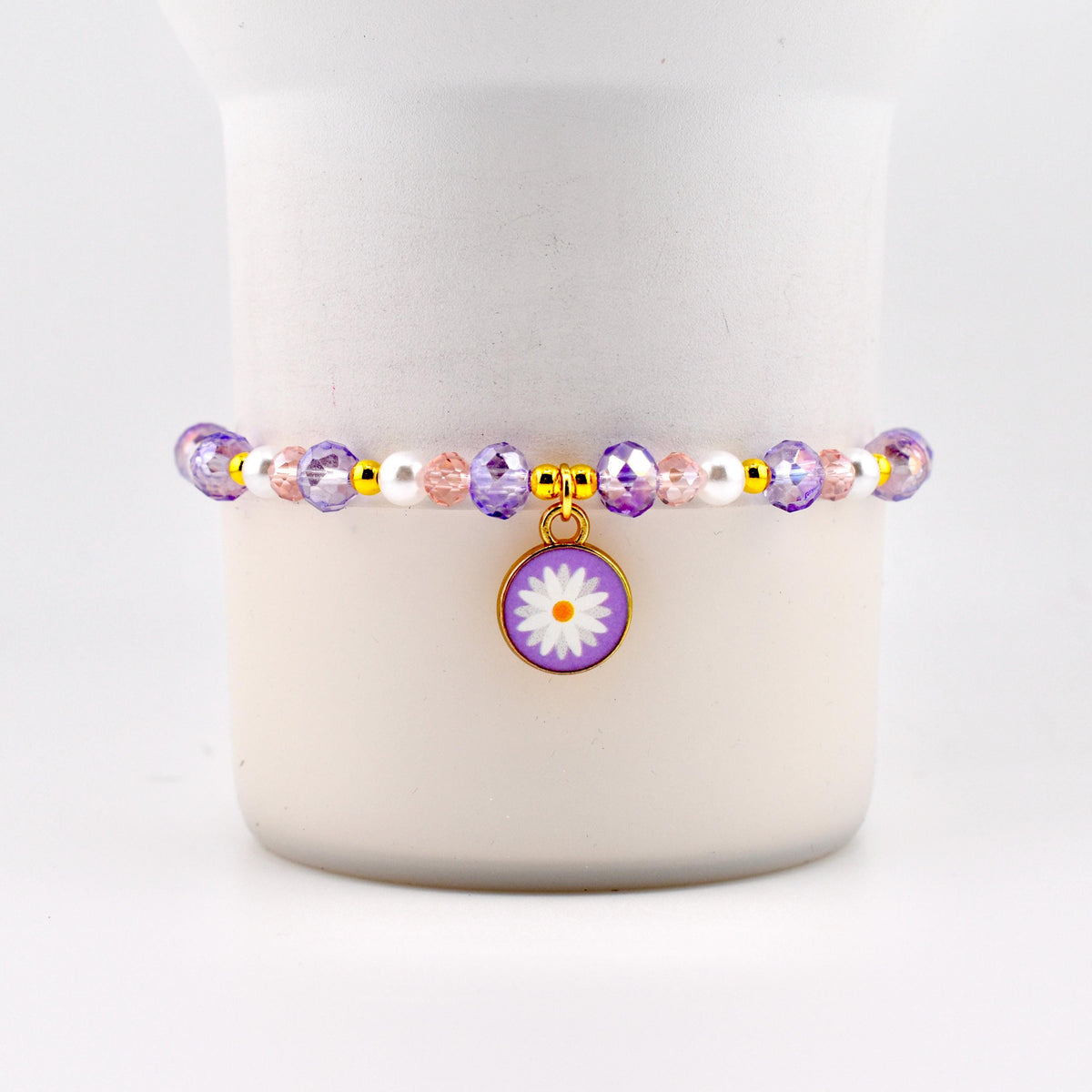 A beaded bracelet featuring a daisy charm. The bracelet has purple, pink, and white beads with gold accents. The daisy charm is purple with a white flower and a gold border. A delicate piece of jewelry.