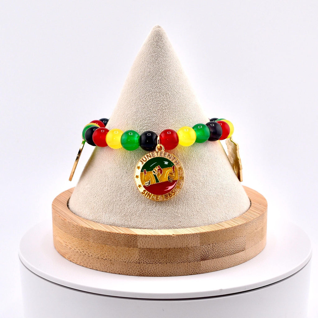 A beaded bracelet with red, yellow, green, and black beads. The bracelet features a gold-colored charm with the words "Juneteenth Since 1865" and a graphic of people raising their hands. The bracelet is displayed on a white cone-shaped stand.