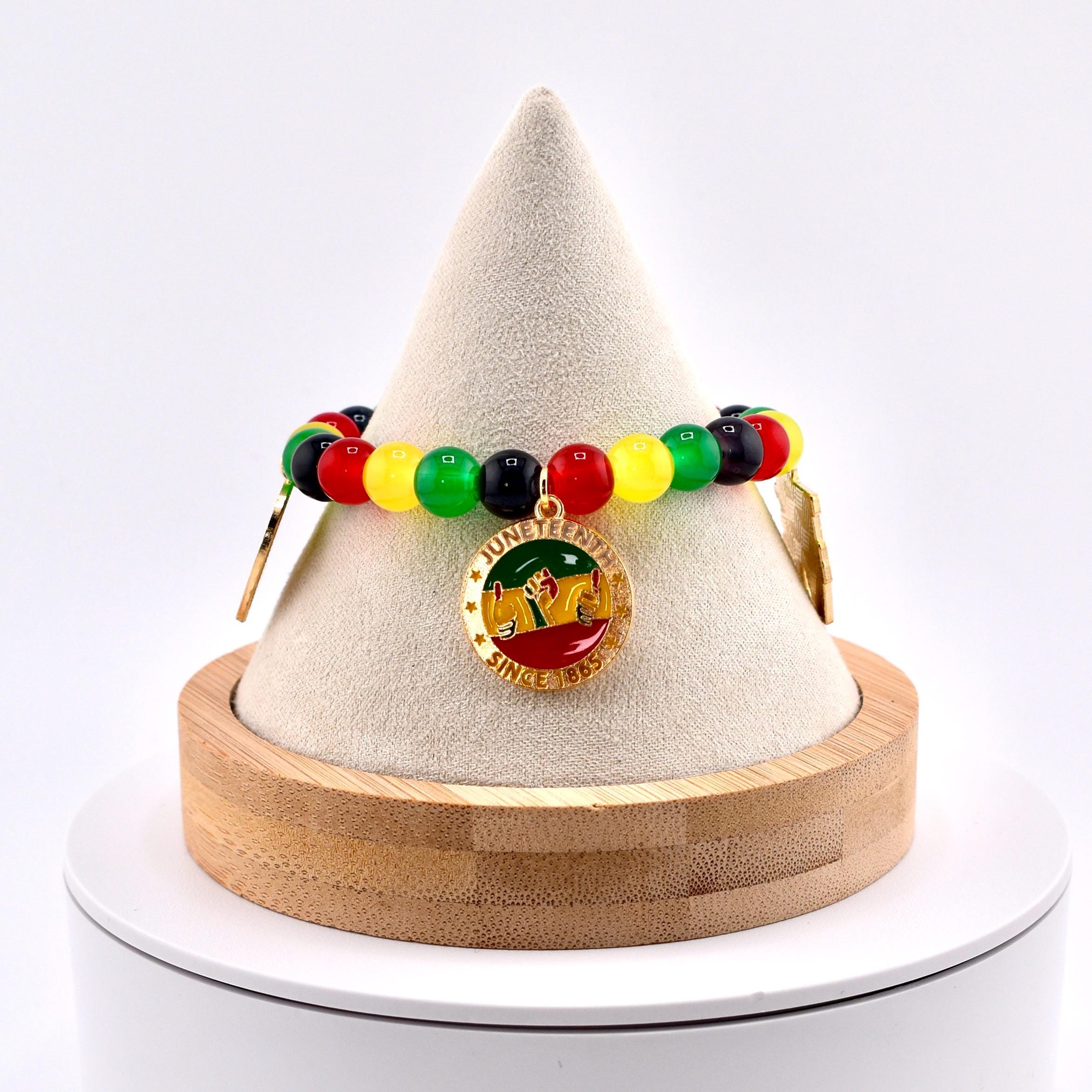 A beaded bracelet with red, yellow, green, and black beads. The bracelet features a gold-colored charm with the words "Juneteenth Since 1865" and a graphic of people raising their hands. The bracelet is displayed on a white cone-shaped stand.