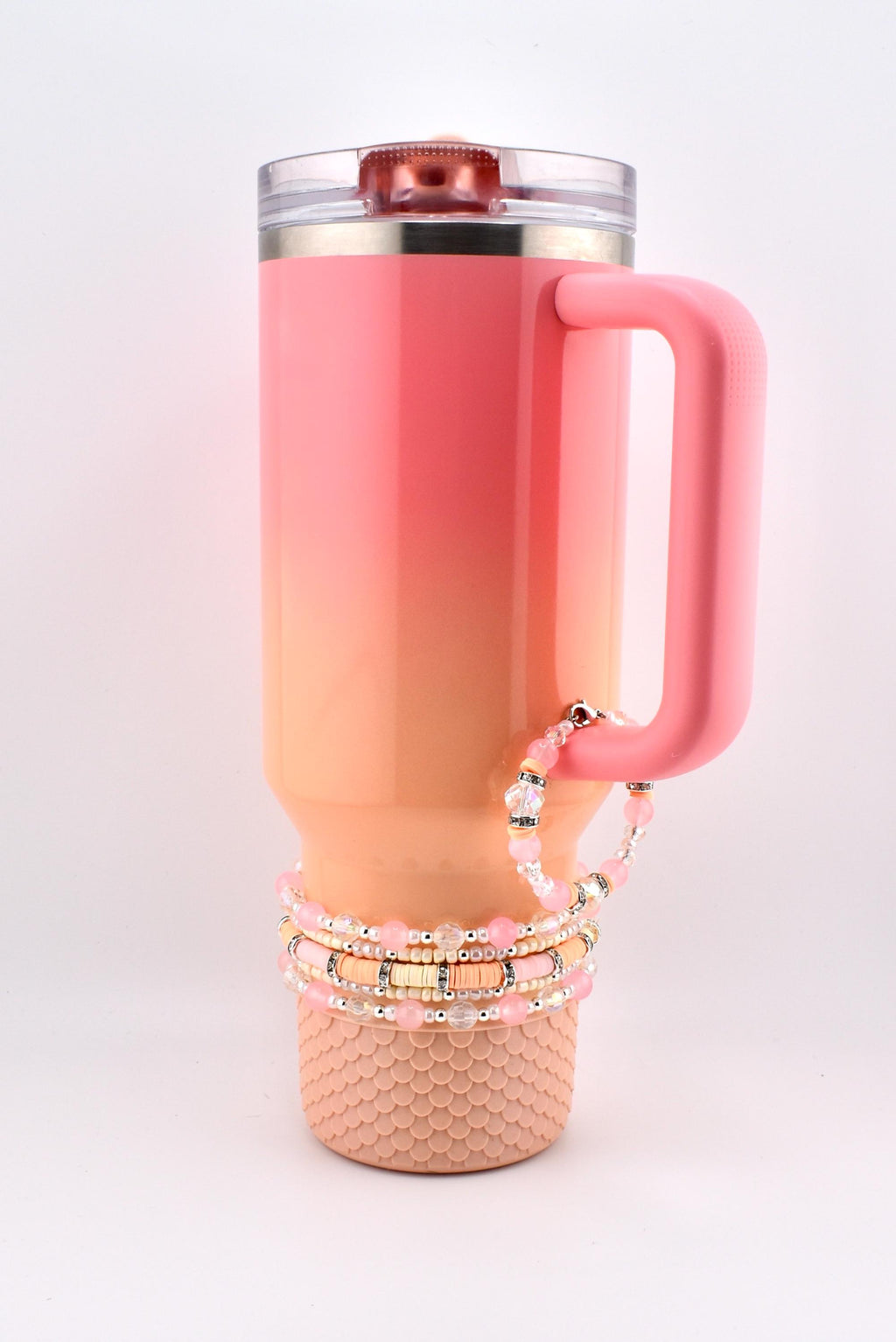 A pink and orange ombre insulated tumbler with a handle, topped with a clear lid. Several pink beaded bracelets are looped around the handle and base of the tumbler. The base has a textured, scale-like design.