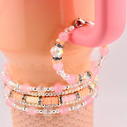 A stack of beaded bracelets in shades of pink, peach, and clear. The bracelets feature a variety of beads, including round, faceted, and disc-shaped beads, with silver accents. The bracelets are displayed on a pink background.