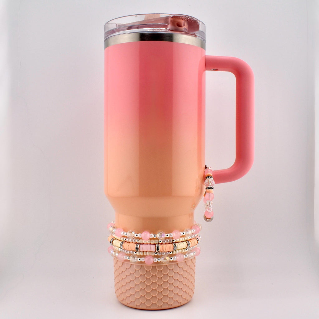 A pink and peach ombre insulated tumbler with a handle and a clear lid. The tumbler is adorned with a stack of beaded bracelets in shades of pink, peach, and gold. The bottom of the tumbler has a textured, scale-like design.