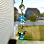 A decorative suncatcher with a clear, faceted crystal at the top, connected to a series of clear and blue beads. A small angel charm with silver wings and a blue crystal body hangs at the bottom. The suncatcher is suspended in front of a window.