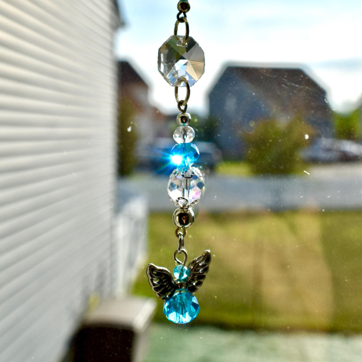 A decorative suncatcher with a clear, faceted crystal at the top, connected to a series of clear and blue beads. A small angel charm with silver wings and a blue crystal body hangs at the bottom. The suncatcher is suspended in front of a window.