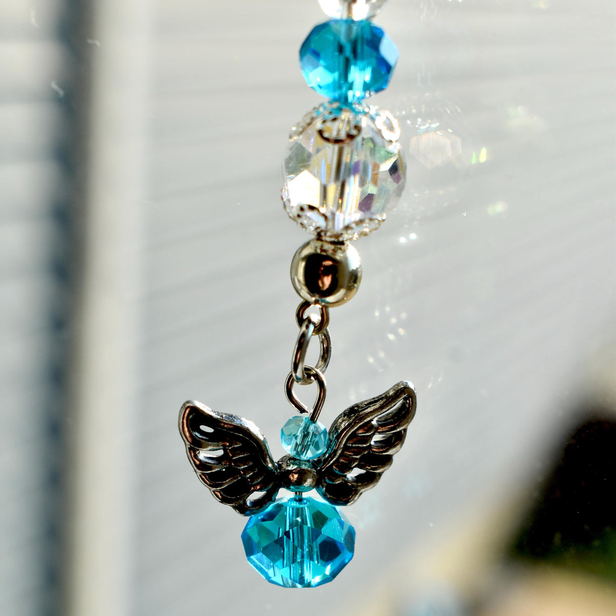 A silver angel charm with detailed wings and a faceted blue crystal body. The charm is suspended from a chain with clear and blue crystal beads. The beads are of varying sizes and shapes.
