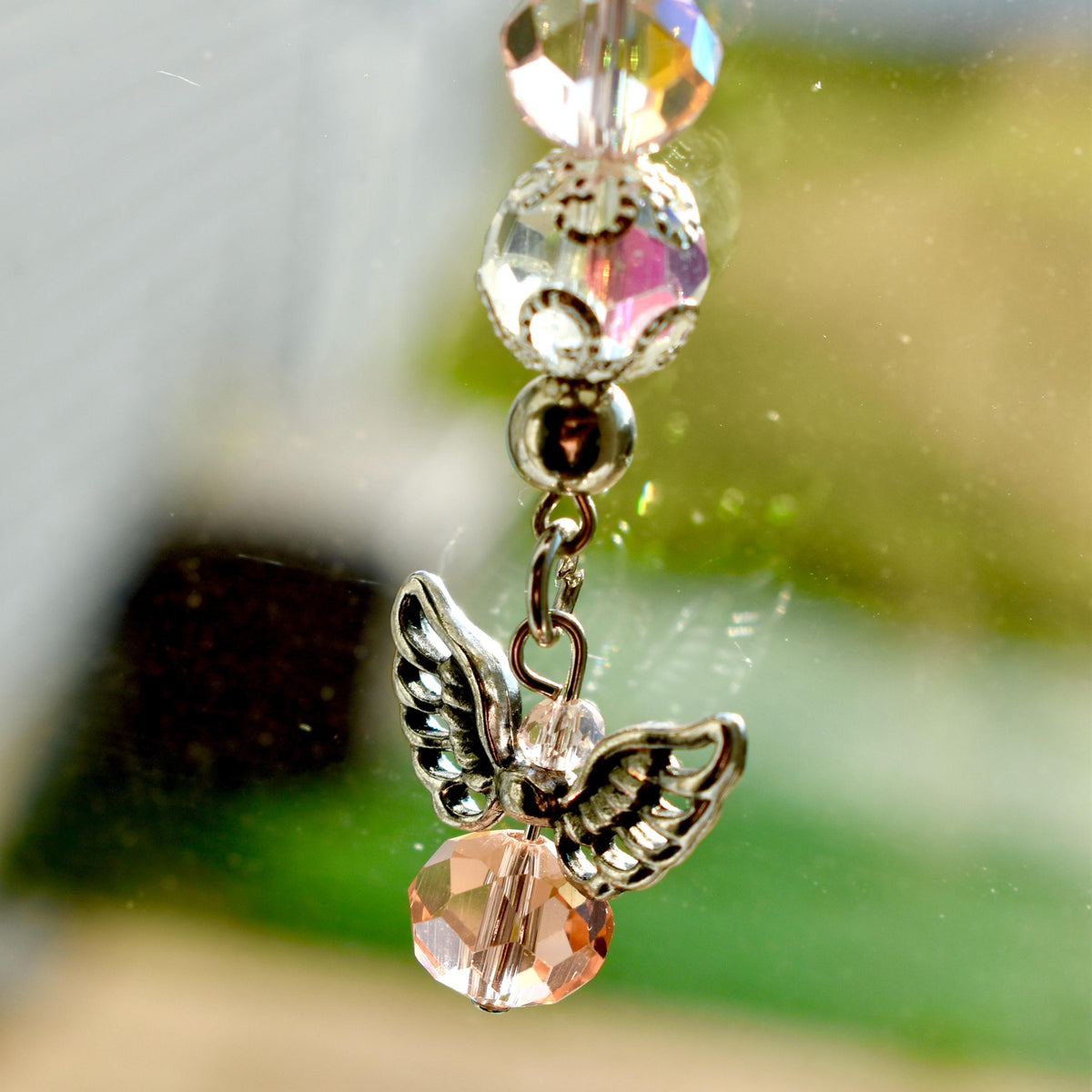 A close-up of a decorative angel charm. The charm features a silver-colored angel with detailed wings, a faceted peach-colored bead, and clear crystal beads. The background is blurred with green and white hues.