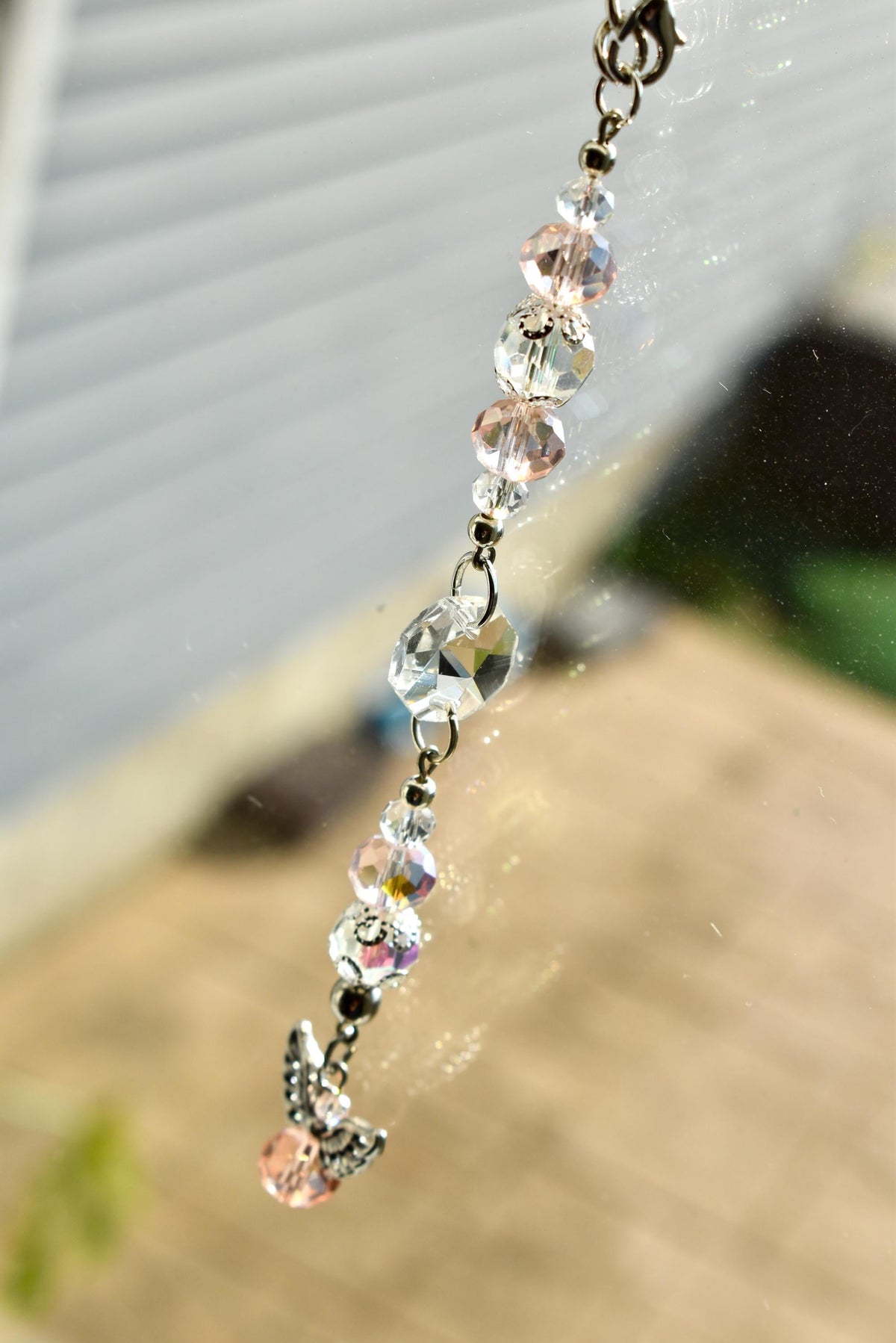 A beaded charm with clear and pink crystal beads and a small silver angel charm. The charm is strung on a silver chain and is hanging in front of a blurred background.
