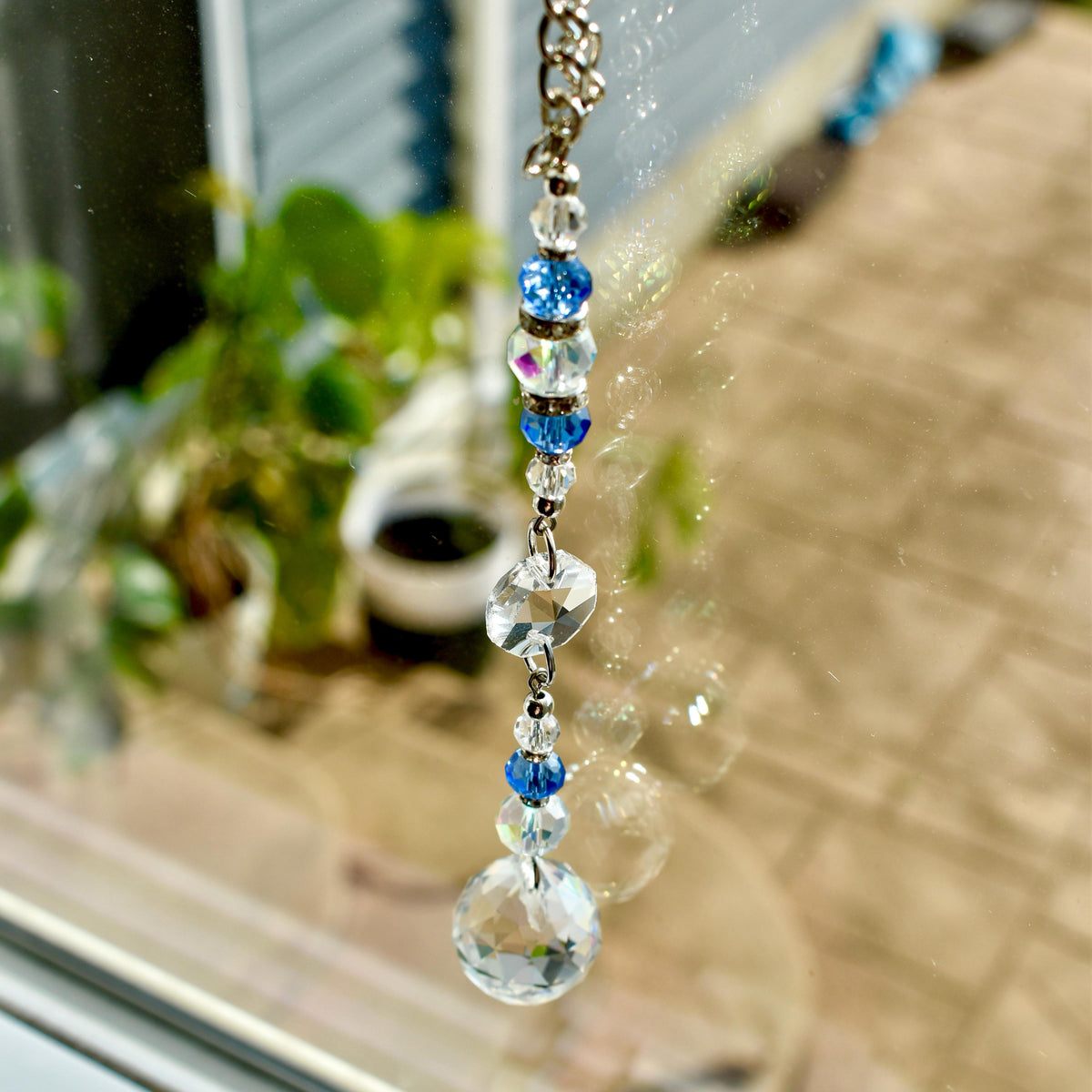 A decorative suncatcher with a silver chain, featuring clear and blue crystal beads. The suncatcher has a large, round crystal at the bottom and a faceted crystal above it. The beads are strung together, creating a sparkling effect.