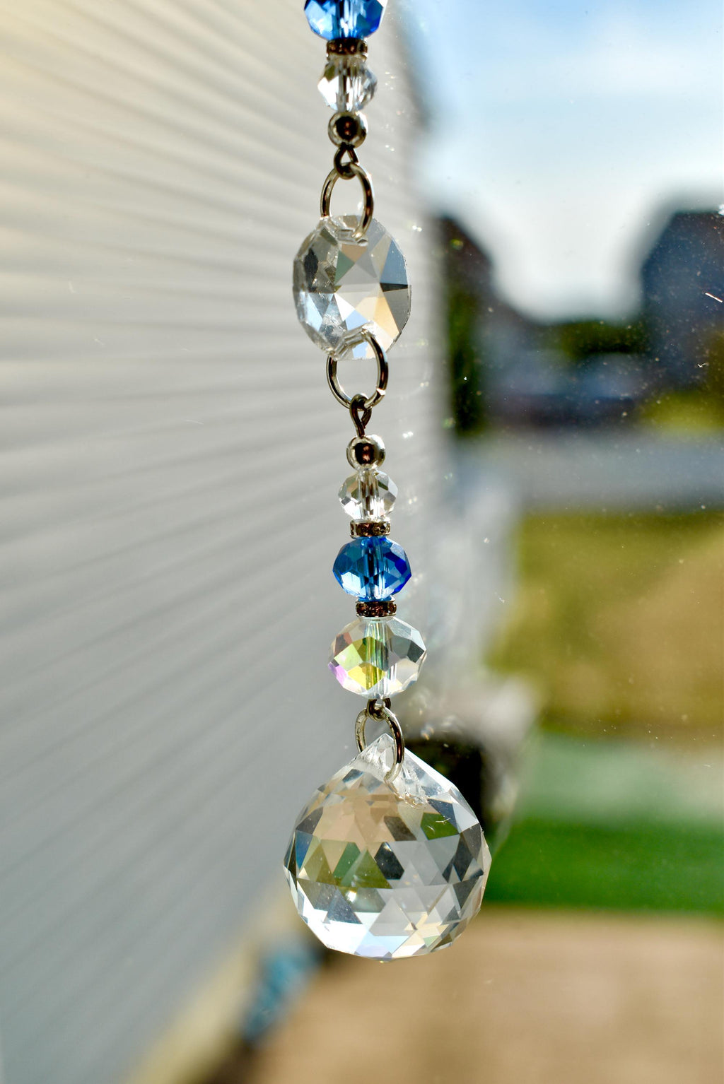 A decorative crystal suncatcher with a series of clear and blue faceted beads and a large, teardrop-shaped crystal at the bottom. The beads are connected by silver-colored metal rings.