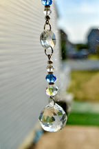 A decorative crystal suncatcher with a series of clear and blue faceted beads and a large, teardrop-shaped crystal at the bottom. The beads are connected by silver-colored metal rings.