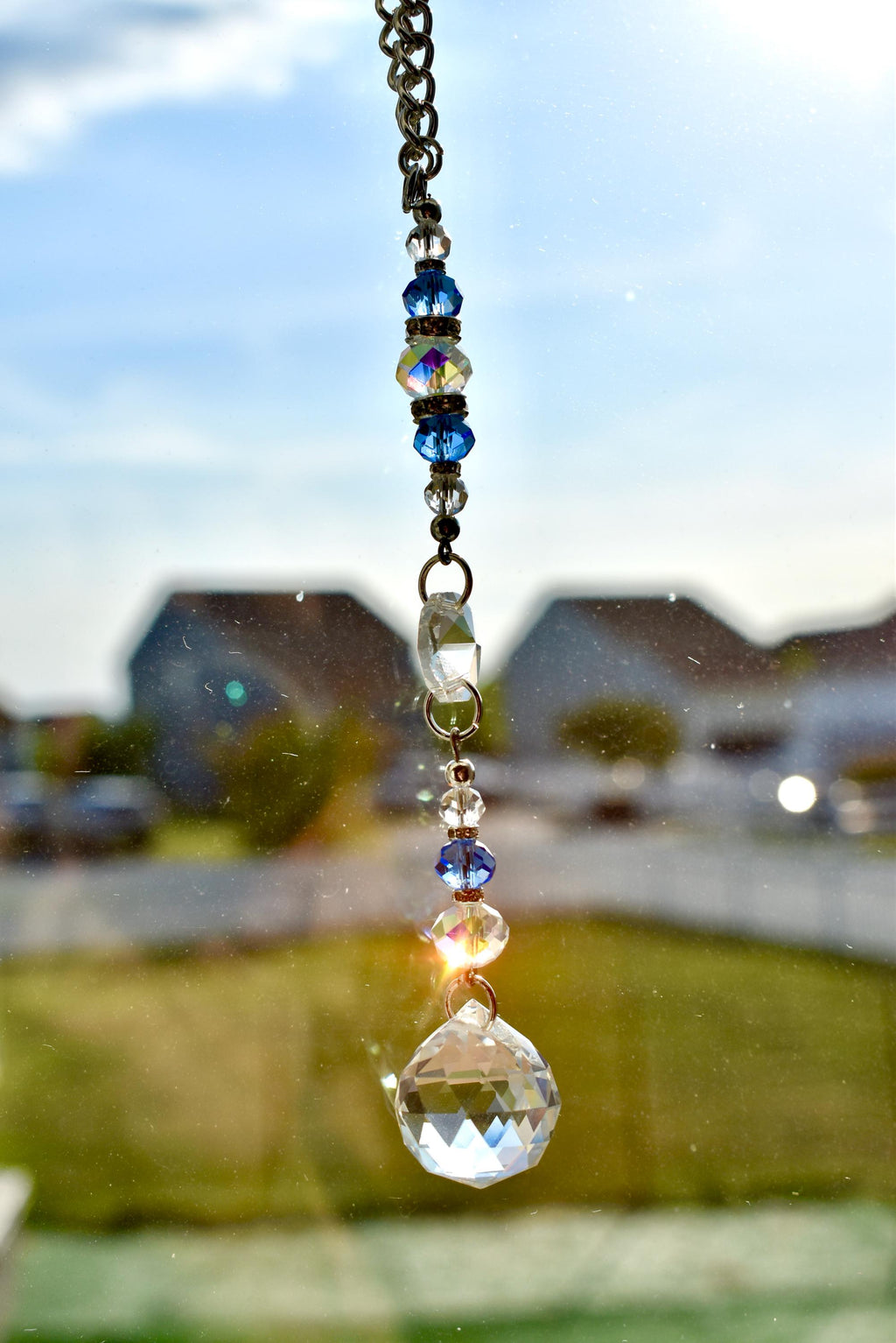 A decorative suncatcher with a silver chain and a series of blue and clear crystal beads. The suncatcher culminates in a large, faceted crystal ball. The background shows a blurred view of a residential area.