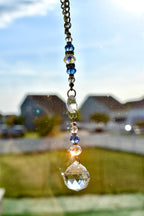 A decorative suncatcher with a silver chain and a series of blue and clear crystal beads. The suncatcher culminates in a large, faceted crystal ball. The background shows a blurred view of a residential area.
