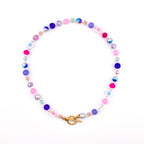 A colorful beaded necklace featuring a variety of beads in shades of pink, purple, blue, and clear. The necklace includes flower-shaped beads and a gold-colored toggle clasp. The beads are strung together to create a circular design.