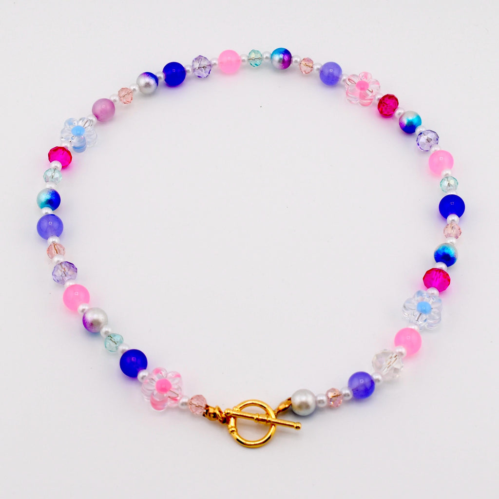 A colorful beaded necklace featuring a variety of beads in shades of pink, blue, purple, and clear. The necklace includes round beads, faceted beads, and flower-shaped beads. It has a gold-colored clasp.