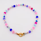 A colorful beaded necklace featuring a variety of beads in shades of pink, blue, purple, and clear. The necklace includes round beads, faceted beads, and flower-shaped beads. It has a gold-colored clasp.