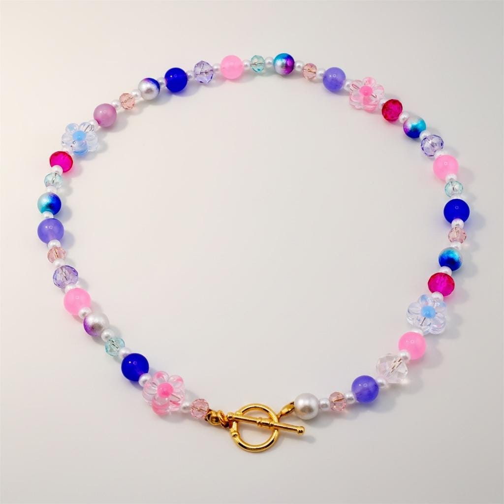 A colorful beaded necklace featuring a variety of beads in shades of pink, blue, purple, and white. The necklace includes flower-shaped beads and a gold-colored clasp. The necklace is designed to be worn around the neck.