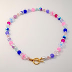 A colorful beaded necklace featuring a variety of beads in shades of pink, blue, purple, and white. The necklace includes flower-shaped beads and a gold-colored clasp. The necklace is designed to be worn around the neck.