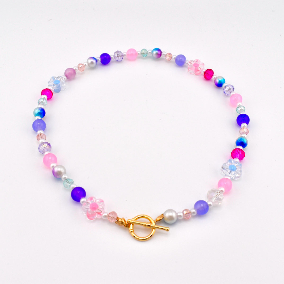 A colorful beaded necklace featuring a variety of beads in shades of pink, blue, purple, and clear. The necklace has a gold-colored toggle clasp and small flower-shaped beads.