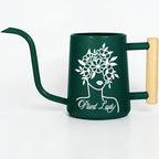 A dark green watering can with a long spout and a wooden handle. The can features a white graphic of a woman's face with a floral crown and the text "Plant Lady".