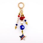 A patriotic keychain with a gold clasp and a dangling charm. The charm features red, white, and blue beads, a blue sphere with a white star, and a star-shaped pendant with a flag design and rhinestones. The word "USA" is spelled out in white letters.
