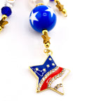 A patriotic charm necklace featuring a star pendant with red, white, and blue enamel and crystal accents. The necklace includes a blue bead with white stars, gold star charms, and blue and white beads.