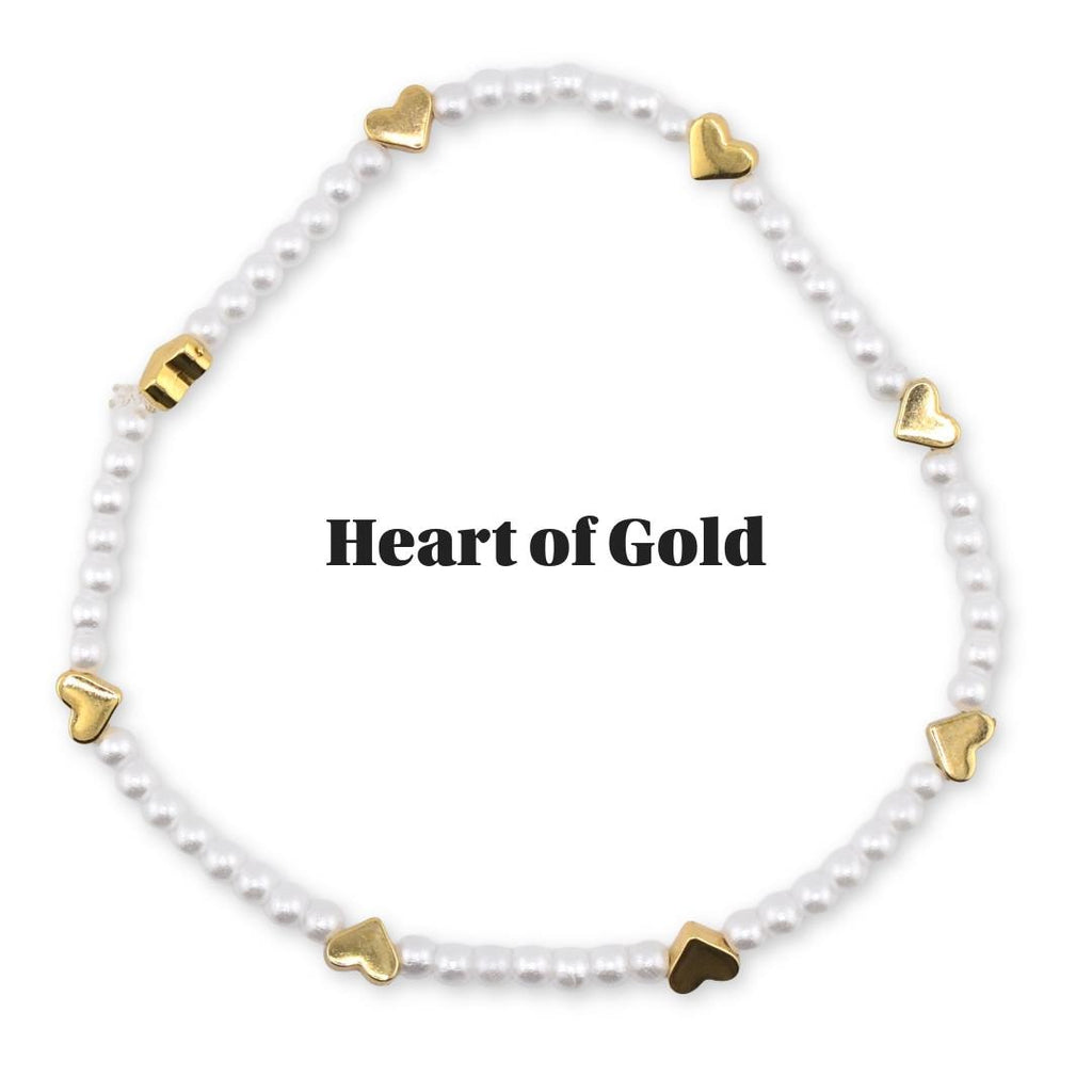 A white beaded bracelet with gold heart-shaped charms. The bracelet is circular and features the text "Heart of Gold" in the center. The bracelet is on a white background.