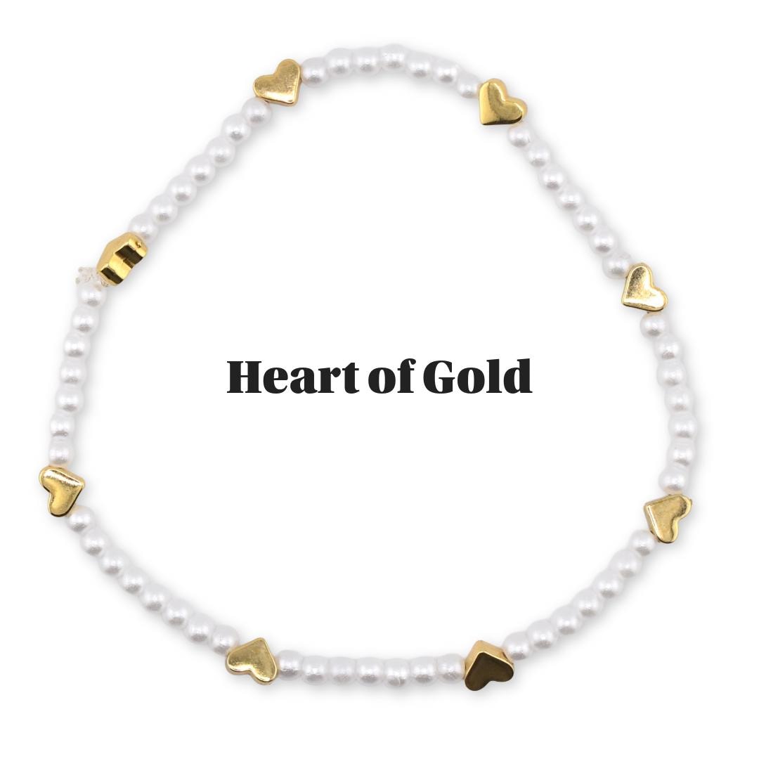 A white beaded bracelet with gold heart-shaped charms. The bracelet is circular and features the text "Heart of Gold" in the center. The bracelet is on a white background.