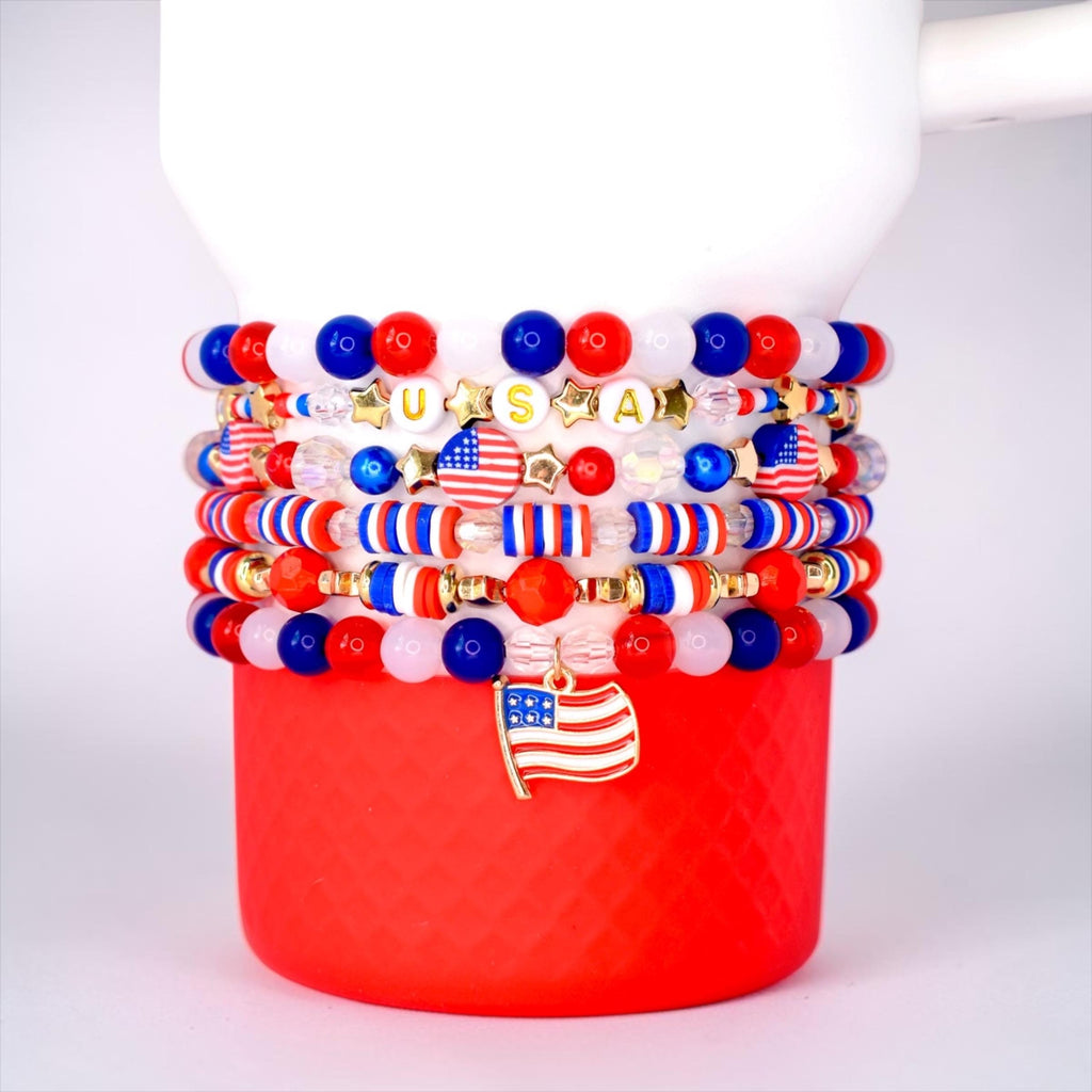 Stack of beaded bracelets in red, white, and blue, with gold accents and a USA charm. The bracelets feature beads of various sizes and shapes, including stars and American flags. The bracelets are stacked on a red cup.