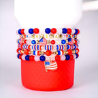 Stack of beaded bracelets in red, white, and blue, with gold accents and a USA charm. The bracelets feature beads of various sizes and shapes, including stars and American flags. The bracelets are stacked on a red cup.