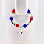 A beaded bracelet featuring red, white, and blue beads, interspersed with silver-colored beads and star-shaped accents. A silver charm with a starburst design hangs from the bracelet. The bracelet is displayed against a white background.