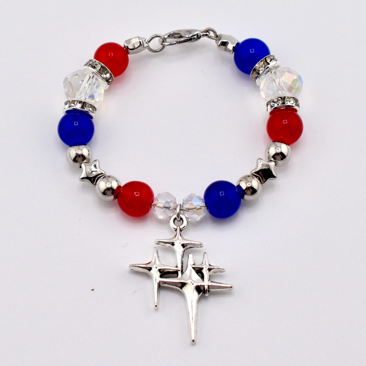 A patriotic bracelet featuring red, white, and blue beads, interspersed with silver star-shaped beads and clear crystal accents. A silver charm with a starburst design hangs from the center. The bracelet is secured with a silver clasp.