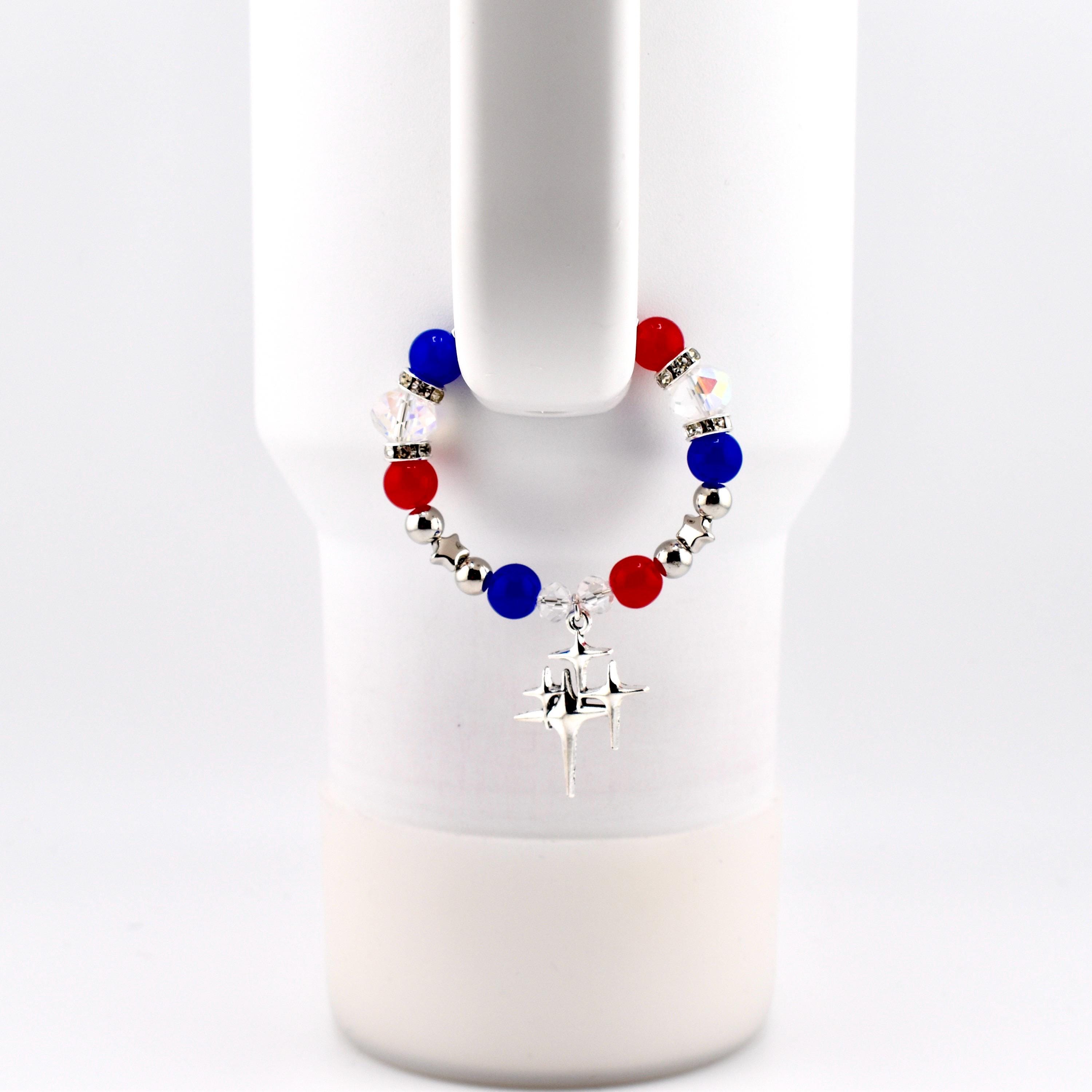 A red, white, and blue beaded tumbler charm bracelet with a silver-colored starburst charm. The handle bracelet is attached to a white mug. The beads are round and interspersed with silver-colored beads and clear crystal beads.