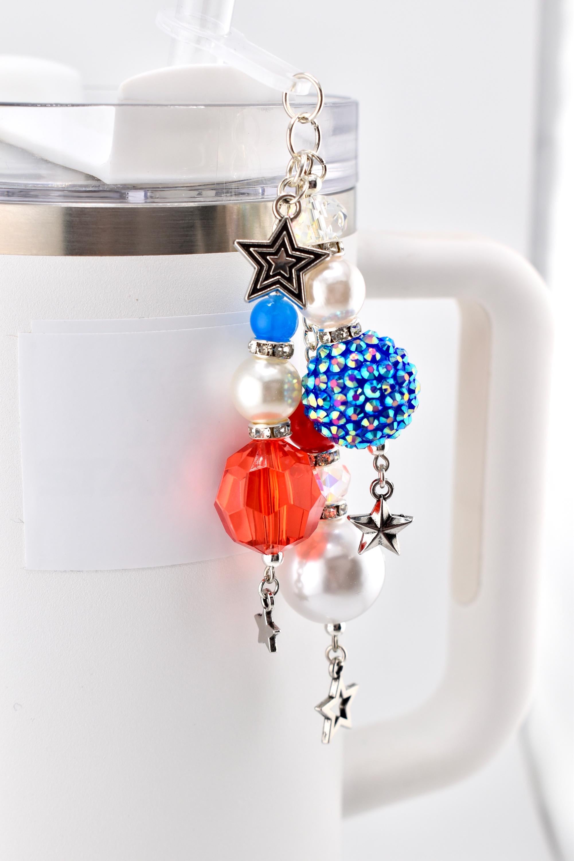 A patriotic-themed charm for a tumbler or water bottle. The charm features a silver chain with red, white, and blue beads, including a faceted red bead, a blue rhinestone ball, and pearl-like beads. Silver star charms dangle from the bottom.