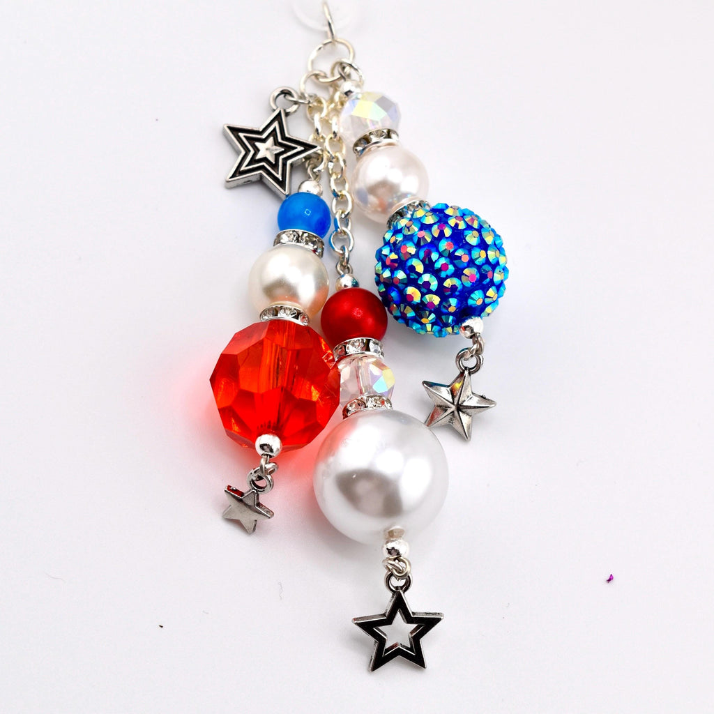 A patriotic charm featuring red, white, and blue beads, including a faceted red bead, a pearlized white bead, and a blue rhinestone-covered bead. Silver star charms dangle from the beads, creating a festive accessory.