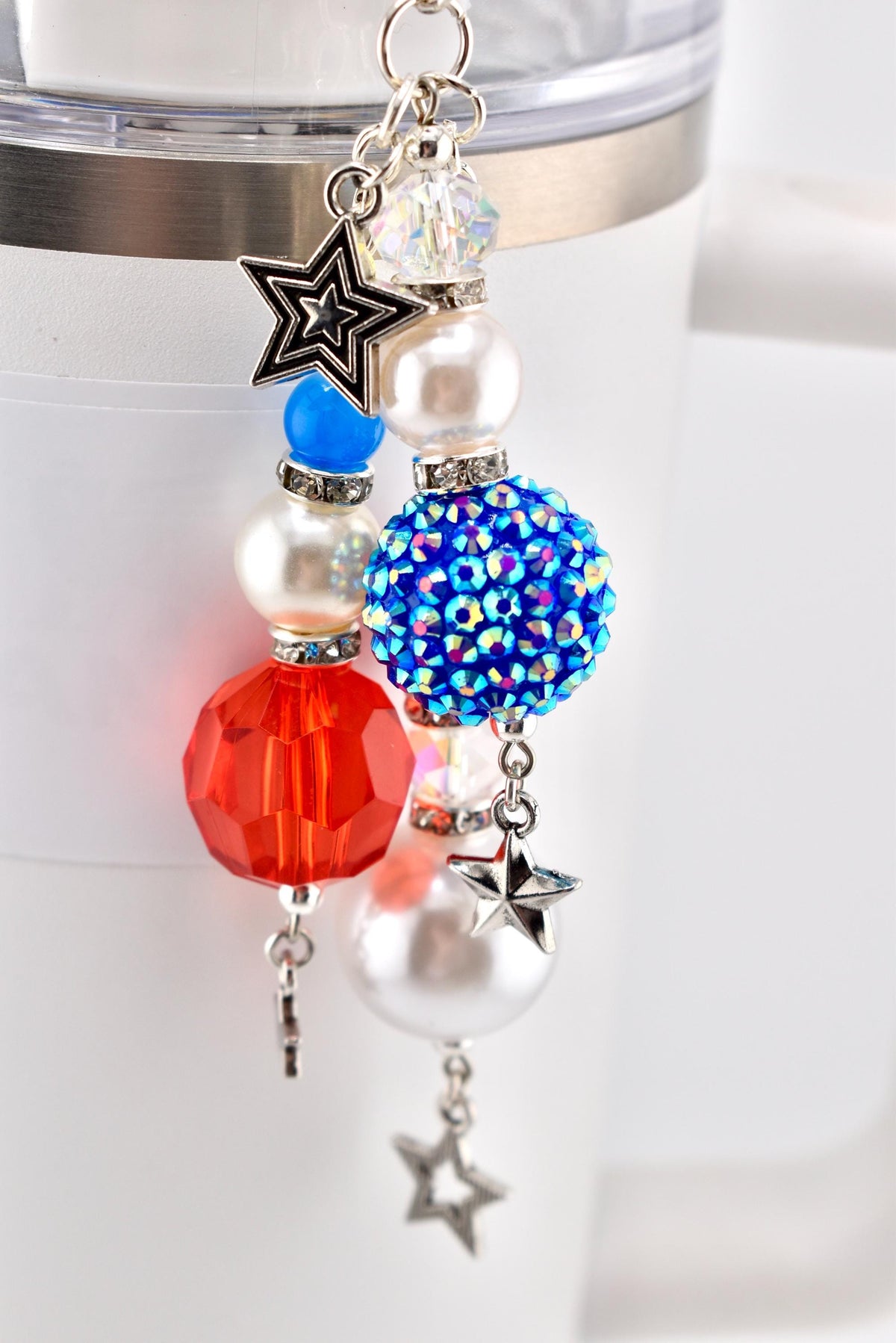 A patriotic-themed charm with red, white, and blue beads and star accents. The charm features a faceted red bead, a blue rhinestone-covered bead, and pearl-like beads, all connected with silver-toned chains and star charms.