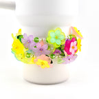 A colorful floral bracelet with yellow, pink, purple, and green flowers. The bracelet is adorned with clear beads and is wrapped around a white cylindrical object. The flowers have a translucent appearance.